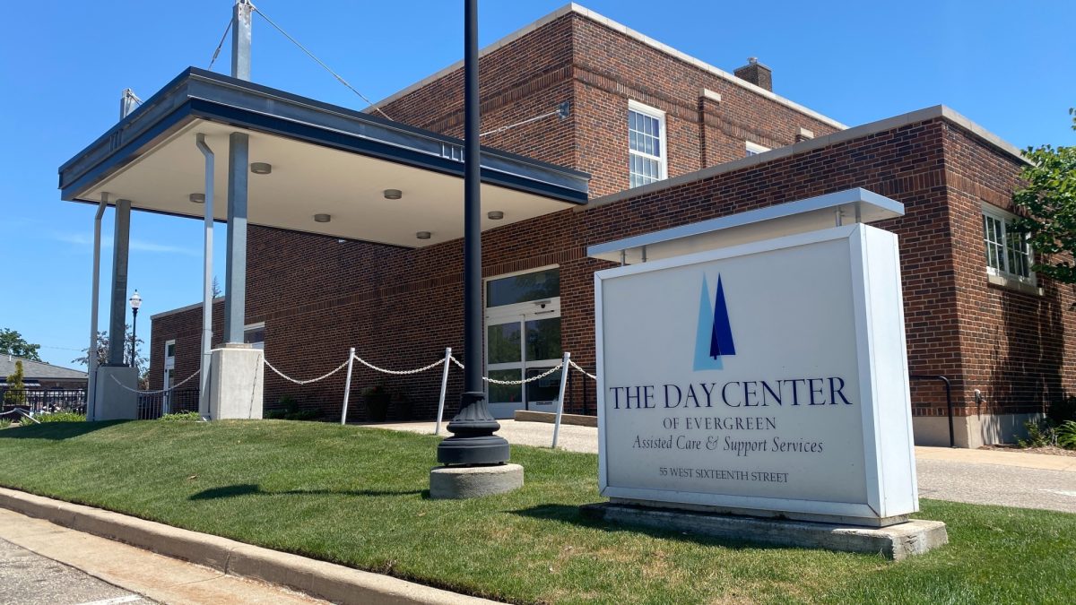 A sign says The Day Center of Evergreen Commons and shows the brick building behind it on a sunny day with a blue sky