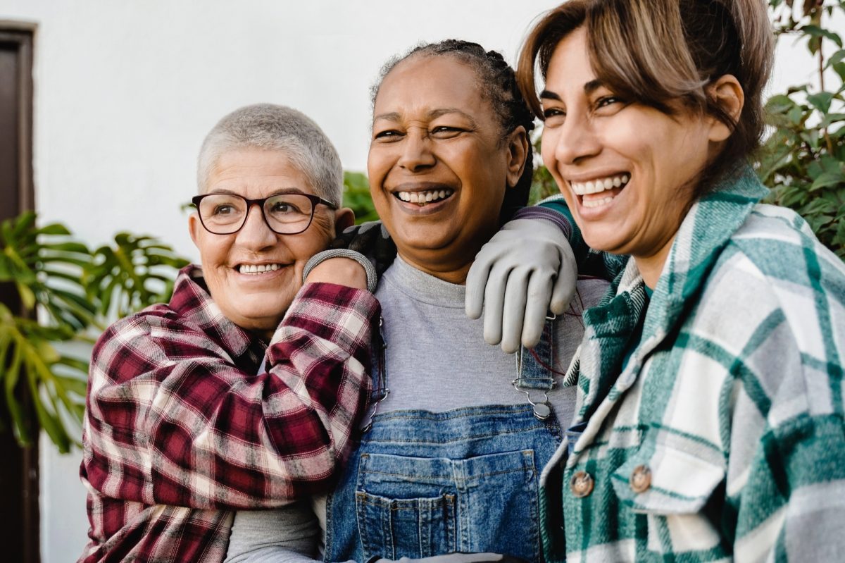 Three adult women smiling looking off into distance wearing gardening gloves overalls and plaid shirts