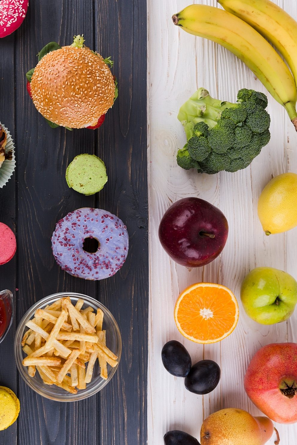 Junk foods such as a burger, donut, and french fries on a black surface next to fresh fruits and vegetables on a white surface.